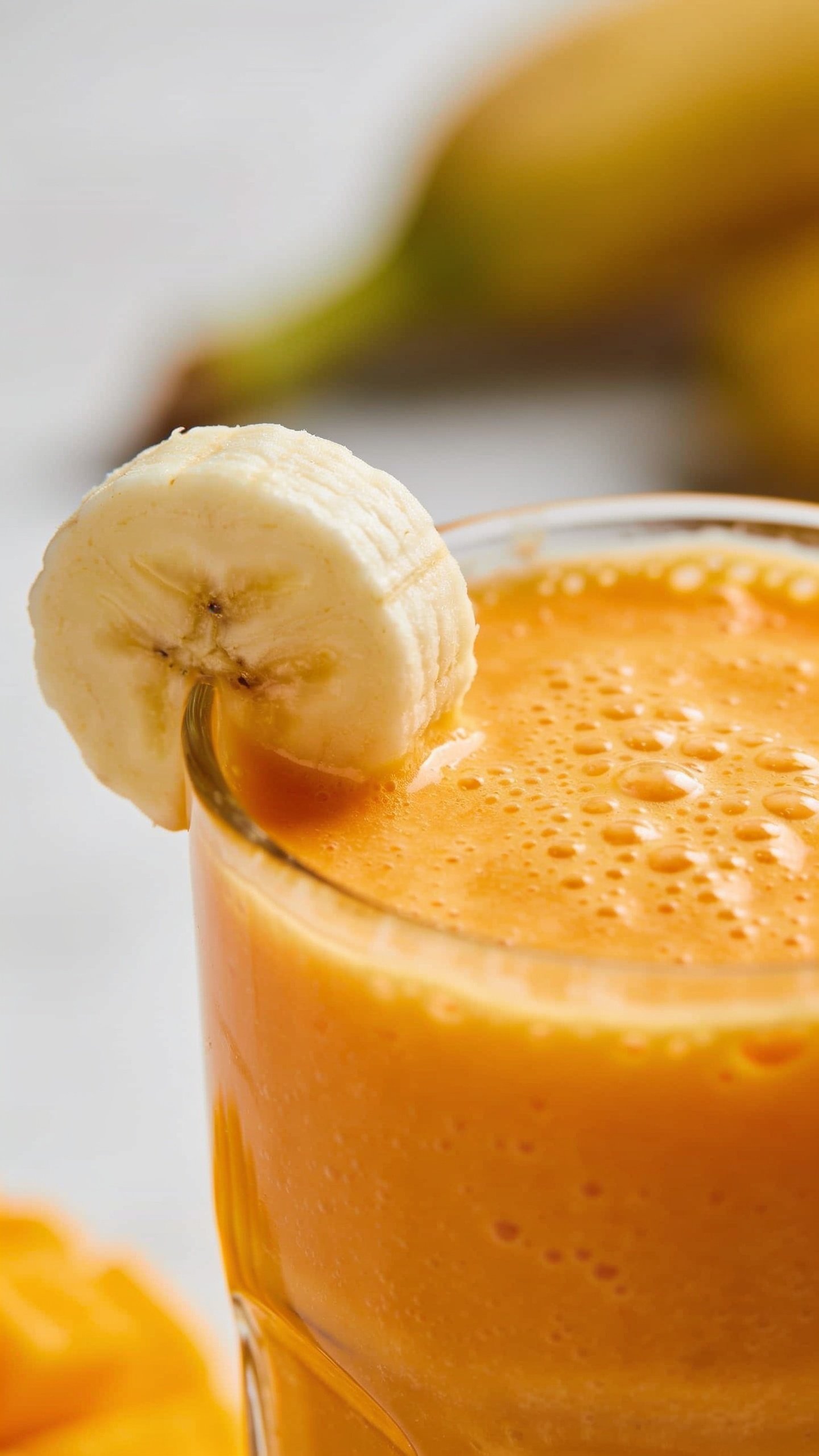 Closeup of whipped mango smoothie surface with banana slice garnish on edge of glass, shallow depth of field